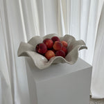 A white sand-colored ceramic wave bowl with a unglazed base, containing peaches as a decorative item, displayed on a pedestal.