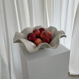 A white sand-colored ceramic wave bowl with a unglazed base, containing peaches as a decorative item, displayed on a pedestal.